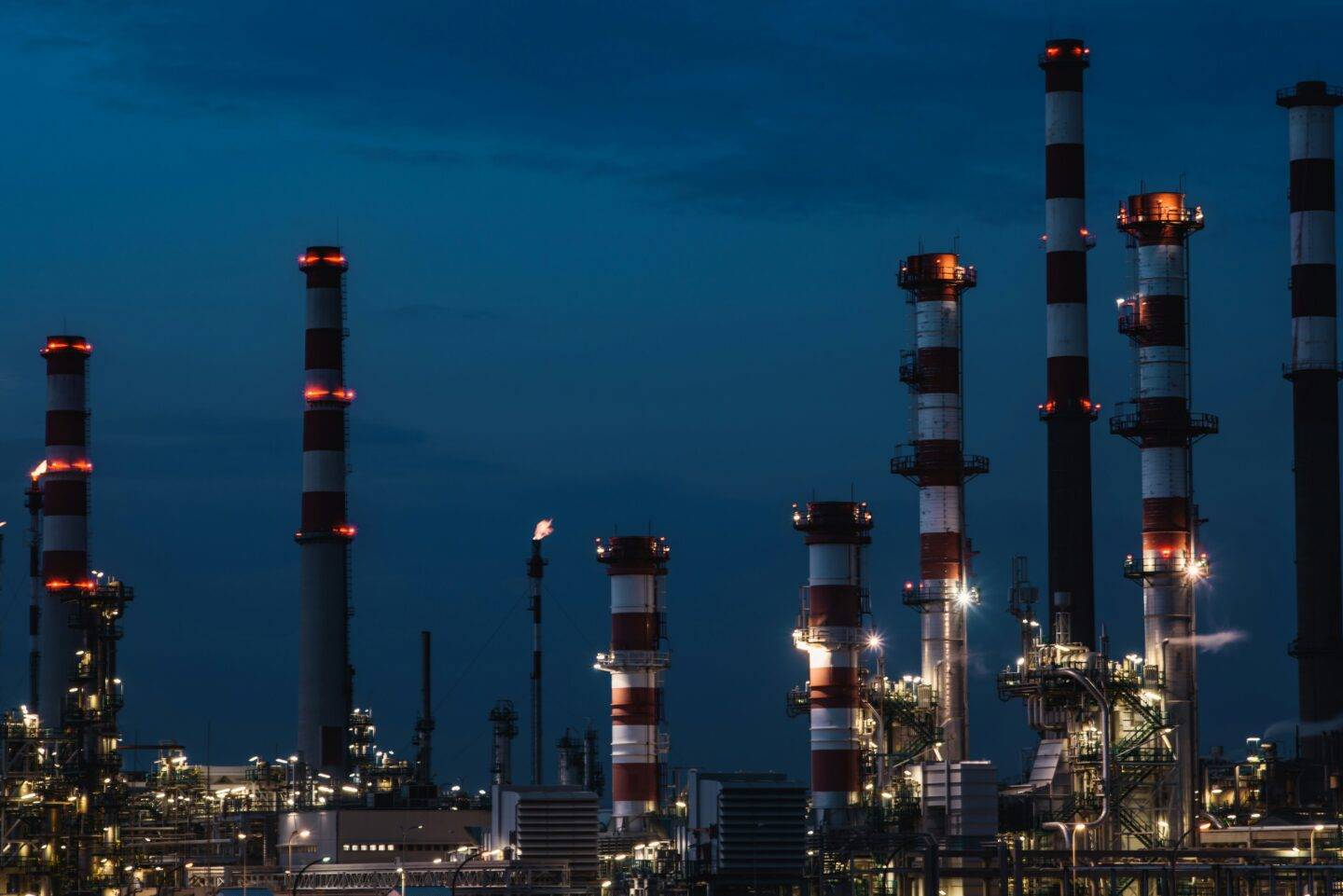 Industrial oil refinery at night, with illuminated red and white striped chimneys and a gas flare against a dark blue sky